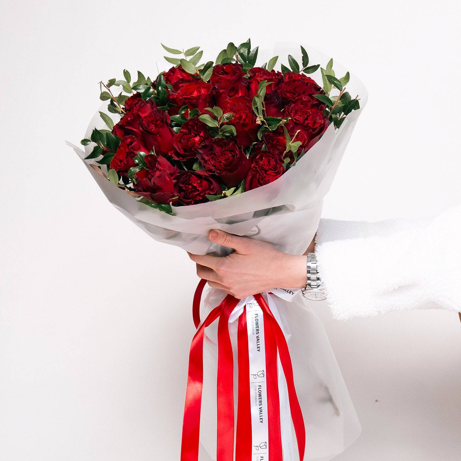 Classic bouquet of deep red roses with greenery in white wrap and red-striped ribbon—held in hand.