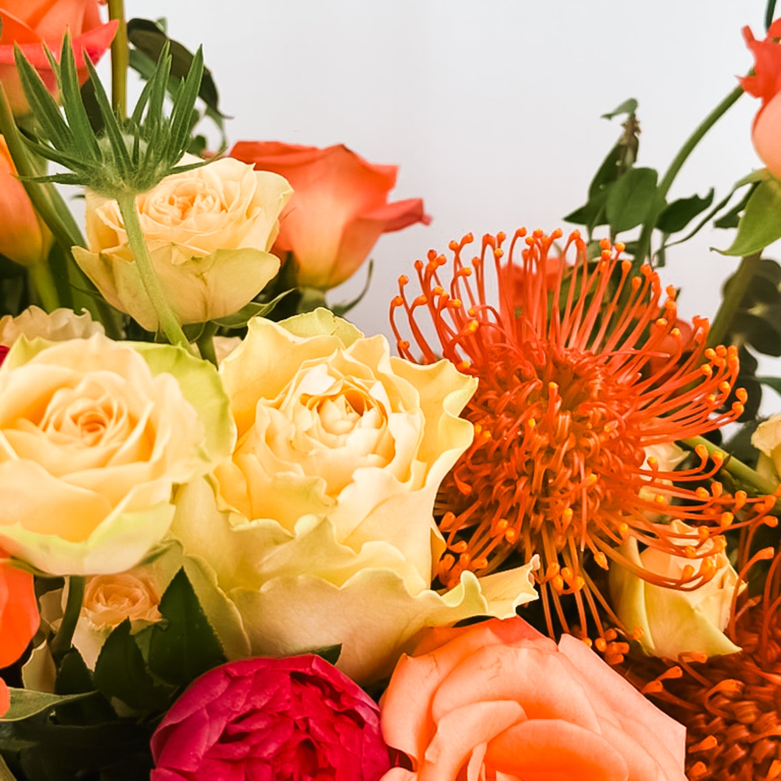 Close-up of orange, peach, and pale yellow roses with bright orange pincushion protea in a luxury floral arrangement.