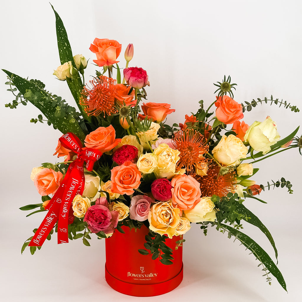 Sunset-colored flower arrangement in a red Flowers Valley box with ribbon, featuring roses, proteas, and greenery.