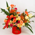 Sunset-colored flower arrangement in a red Flowers Valley box with ribbon, featuring roses, proteas, and greenery.