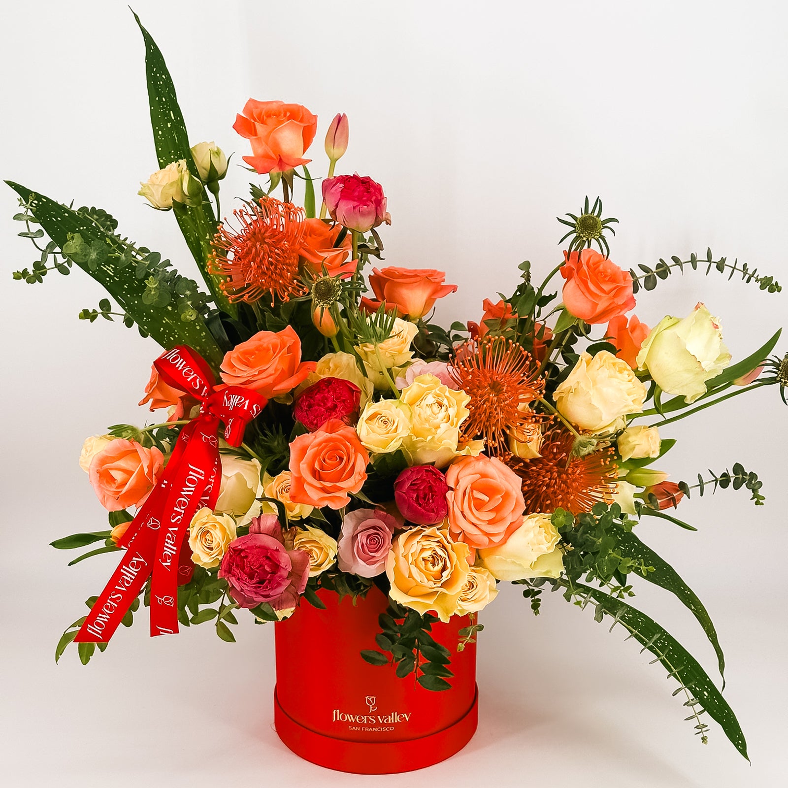 Sunset-colored flower arrangement in a red Flowers Valley box with ribbon, featuring roses, proteas, and greenery.