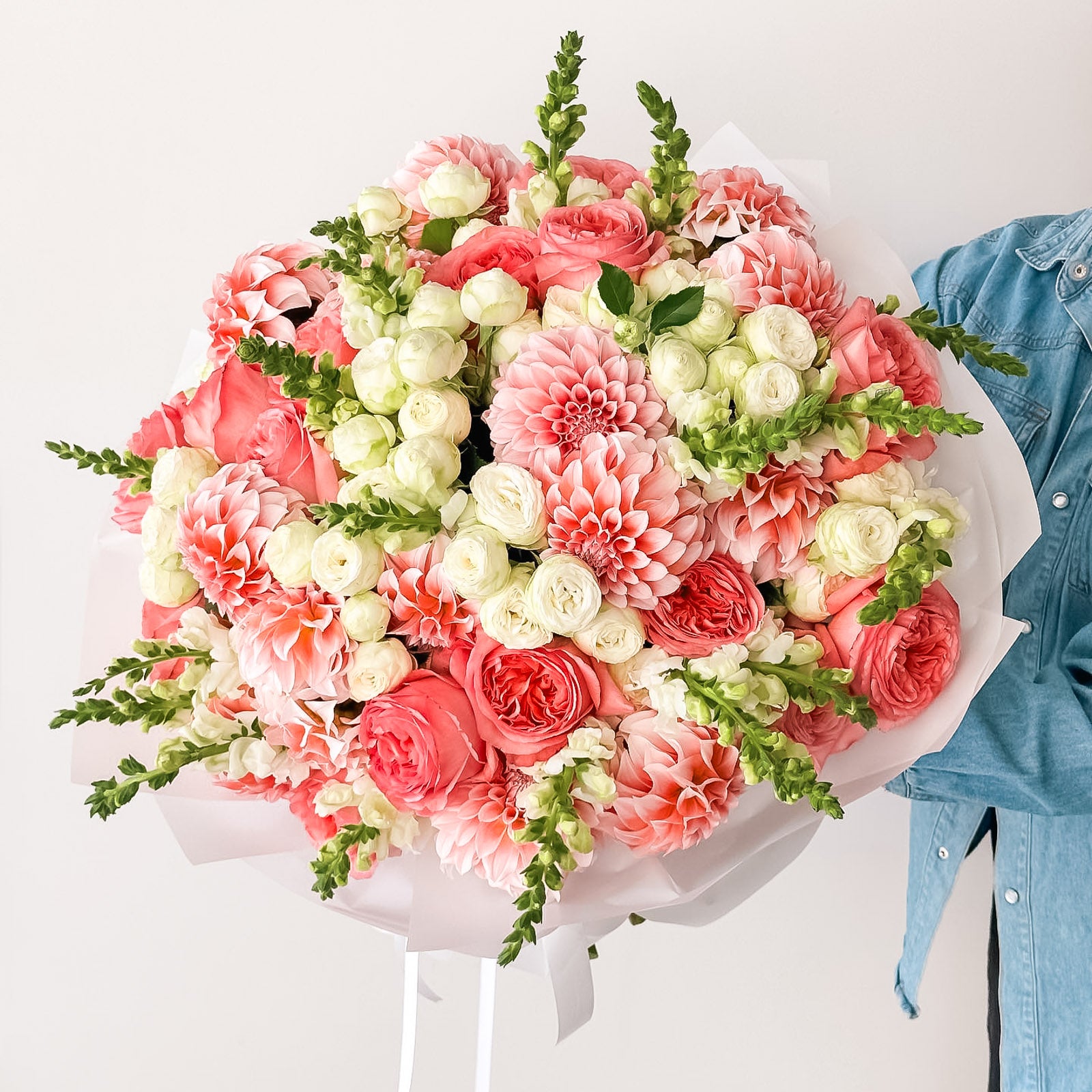 Bouquet of coral dahlias, garden roses, spray roses, and green snapdragons arranged in white wrap—fresh, bright, and modern floral design.