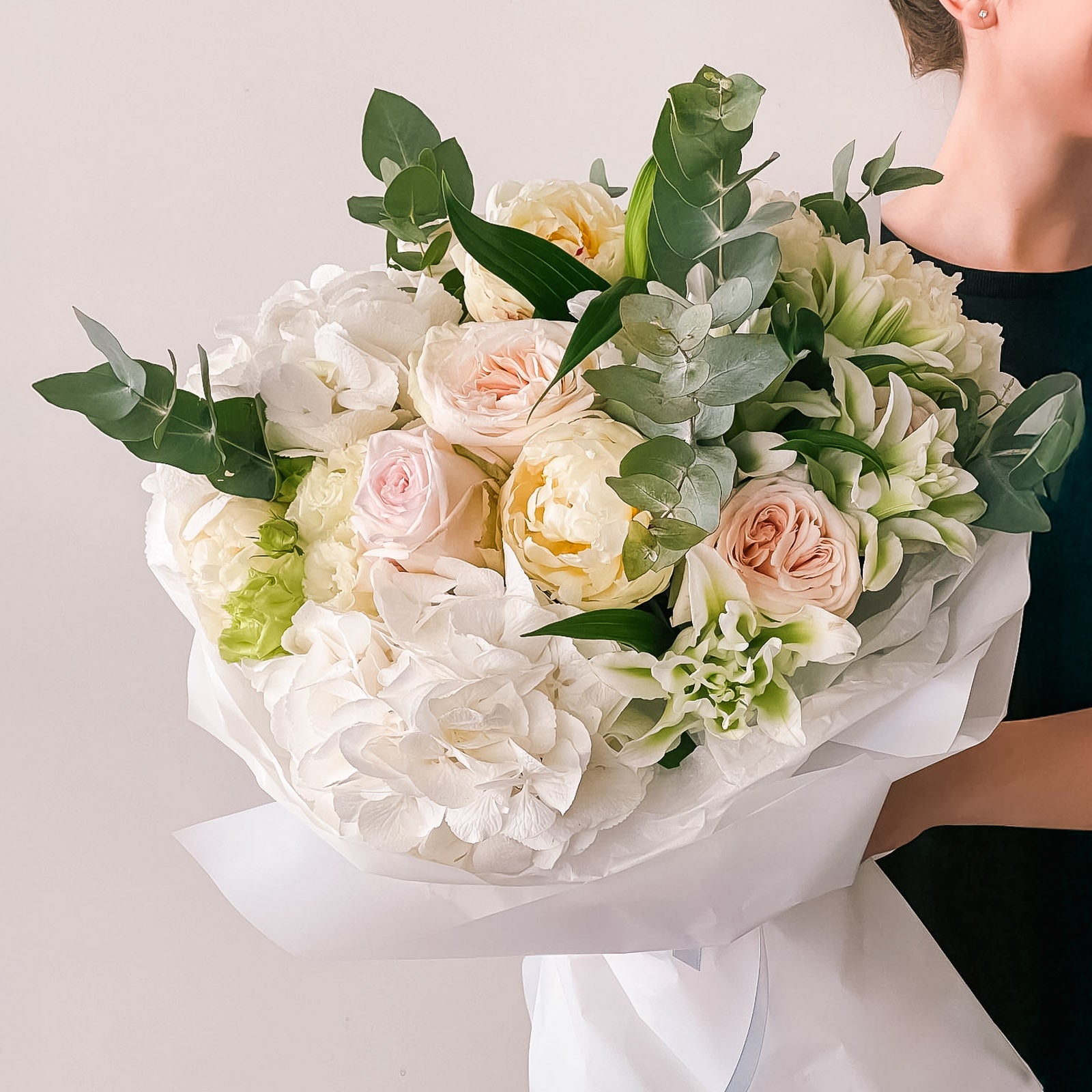 Elegant bouquet of white hydrangeas, ivory peonies, blush roses, lisianthus, and greenery wrapped in crisp white paper—held by a person in black.