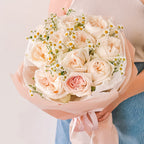 Bouquet of creamy blush garden roses and chamomile wrapped in blush pink paper—held by person in a neutral outfit with satin ribbon detail.