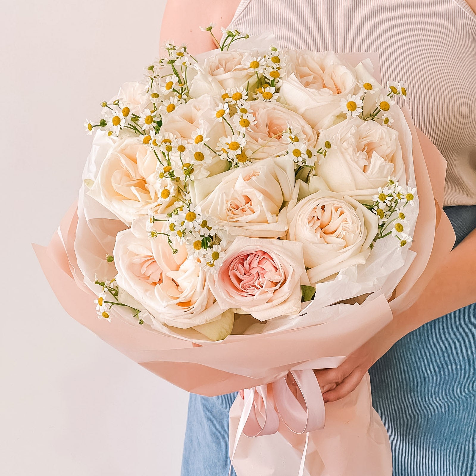 Bouquet of creamy blush garden roses and chamomile wrapped in blush pink paper—held by person in a neutral outfit with satin ribbon detail.