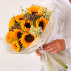 Bouquet of vibrant yellow sunflowers with chamomile and fillers wrapped in white paper—held by a person in white.