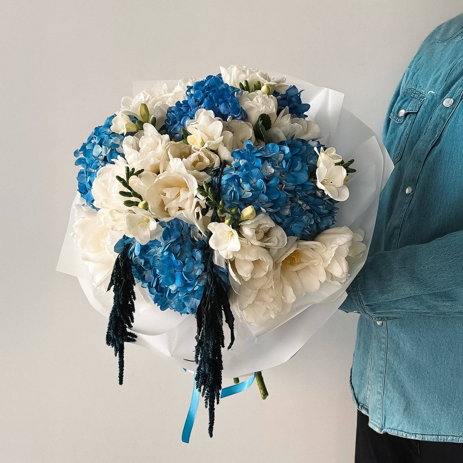 Celeste Bloom bouquet with blue hydrangeas, white lisianthus, and freesias wrapped in white paper