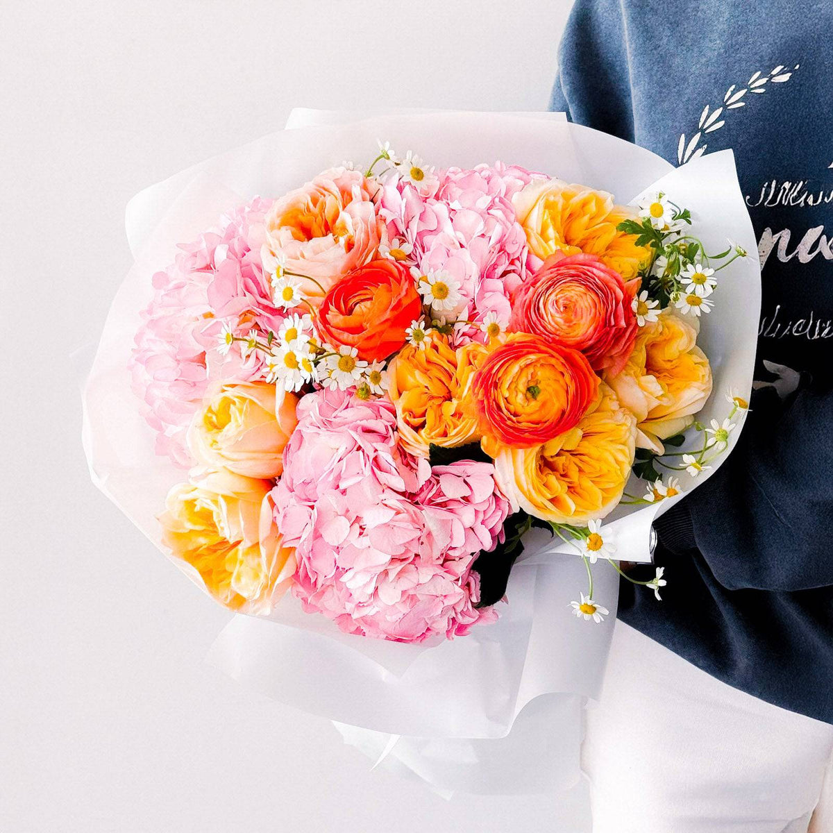 Bright bouquet of orange ranunculus, pink hydrangeas, and cream roses held in white wrap—fresh and vibrant floral design.
