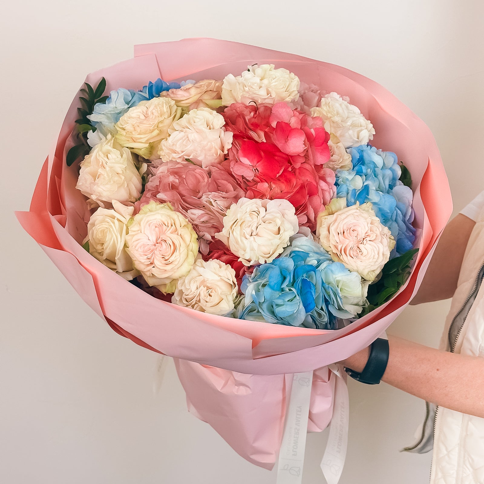 Pastel bouquet of blue hydrangeas, blush roses, and soft pink blooms wrapped in pink paper—held by a person in white.