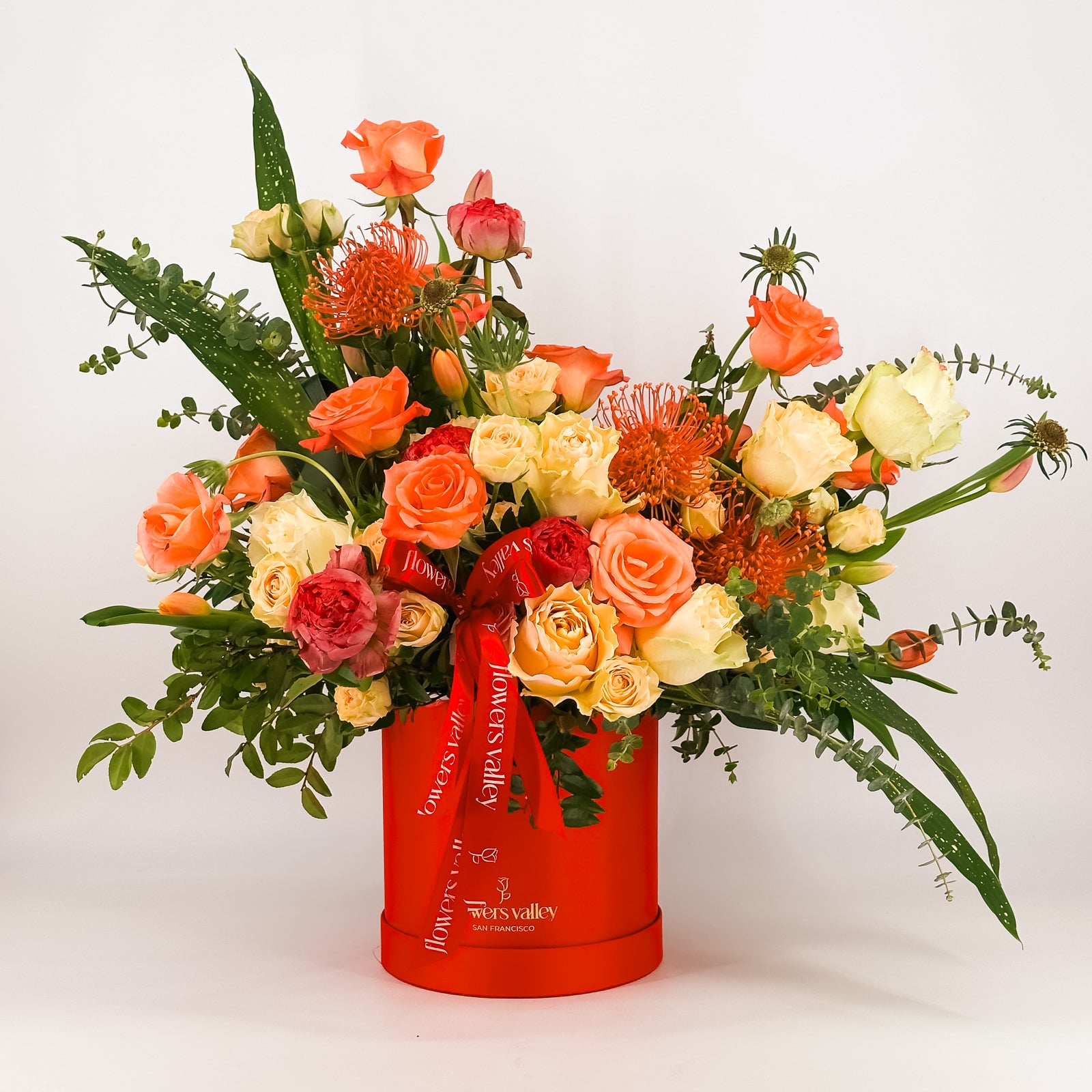 Elegant mixed floral display in coral and yellow tones arranged in red hat box with lush foliage and designer ribbon.