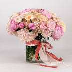 Pastel pink, cream, and peach peonies and roses arranged in a dome shape inside a clear vase with a satin Flowers Valley ribbon.
