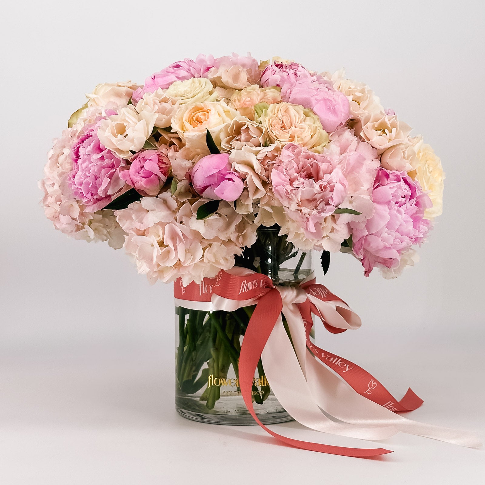 Pastel pink, cream, and peach peonies and roses arranged in a dome shape inside a clear vase with a satin Flowers Valley ribbon.