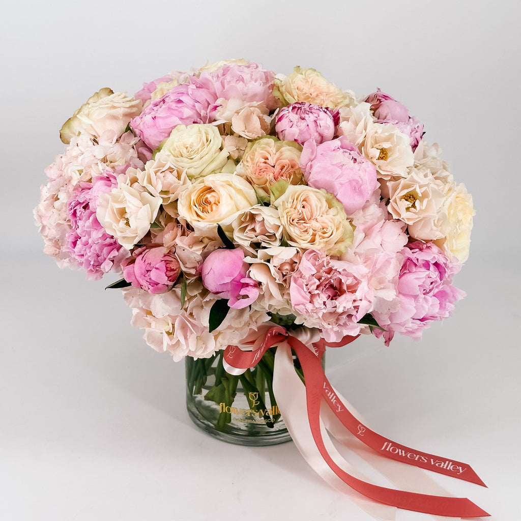Angled view of lush pastel peonies and roses arranged in a clear glass vase, finished with pink and ivory branded ribbons.