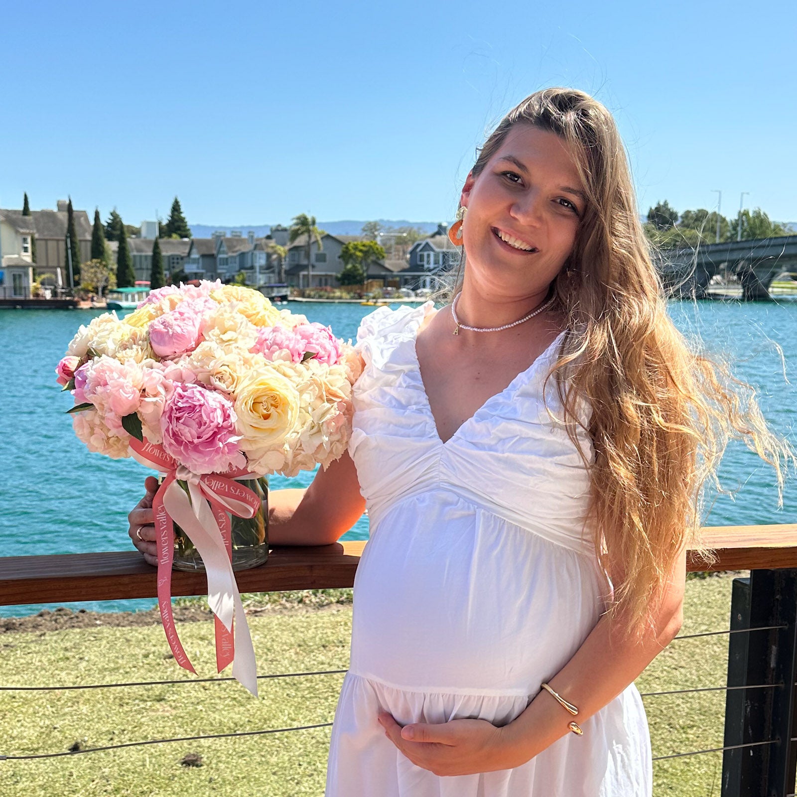 Smiling woman in white dress holding a pastel floral vase arrangement of roses, peonies, and hydrangeas by the waterfront.