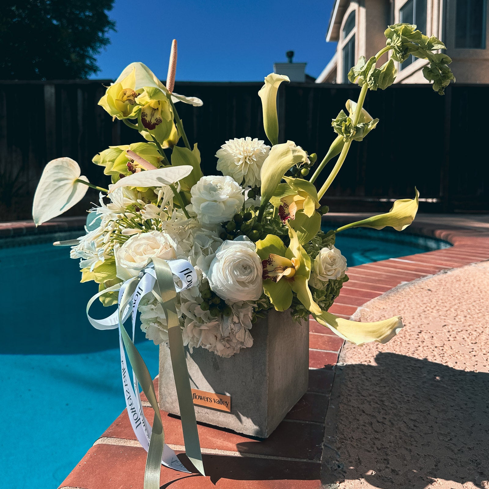 Urban Serenity flower arrangement in concrete vase, featuring white and green blooms, photographed outdoors by a pool for local delivery.